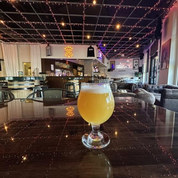 A glass of beer on a reflective table in a brewery with string lights on the ceiling.