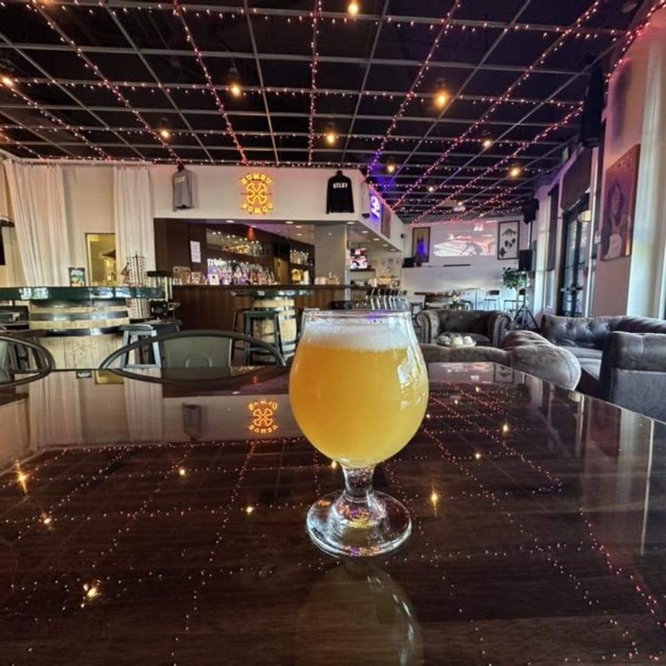 A glass of beer on a reflective table in a brewery with string lights on the ceiling.