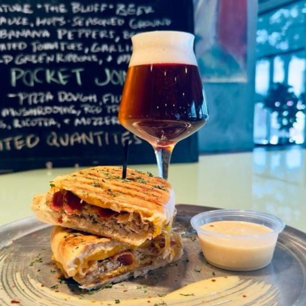 A panini with a side of sauce and a glass of Atlantucky Brewing beer on a metal plate.